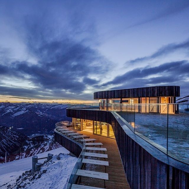 An elegant mountain-top restaurant features outdoor seating with breathtaking panoramic mountain views at dusk.