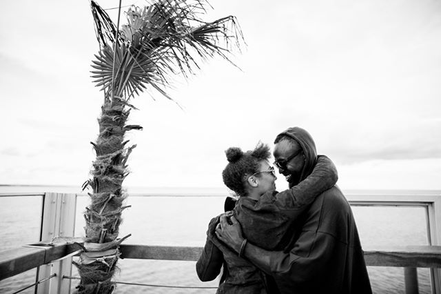 A couple embraces near a palm tree by the coast, captured in a timeless black and white style.