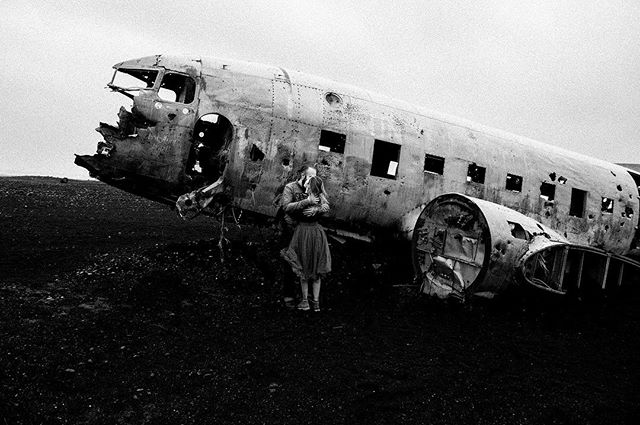 A couple embraces near the wreckage of a plane in a stark, monochrome landscape.