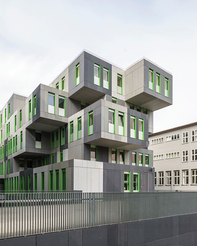 Modern architectural design featuring gray building with green window frames against a cloudy sky in an urban setting.