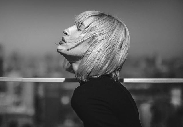 Black and white portrait of a blonde woman looking up with her eyes closed on a rooftop.
