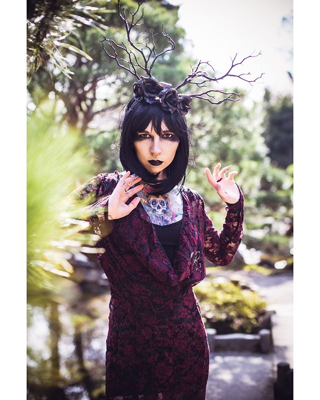 A woman in gothic fashion poses in a garden, showcasing an alternative and mysterious style.