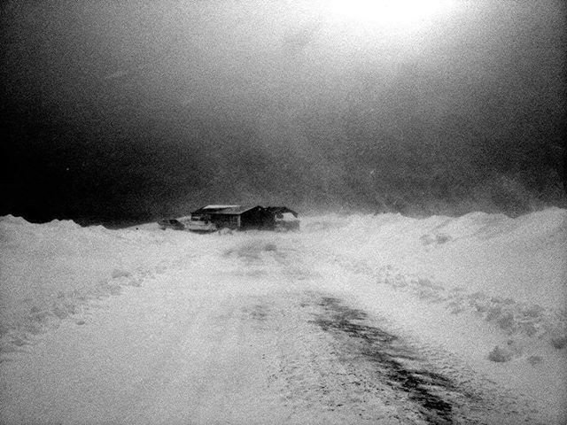 A lone house stands against a stark, snow-covered landscape during a winter storm, rendered in high-contrast black and white.