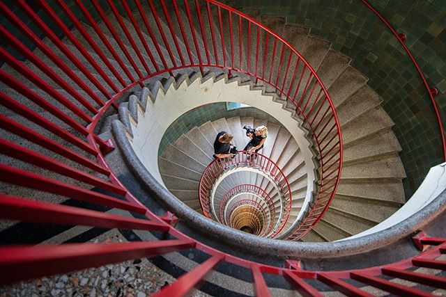 A high-angle shot captures two women on a striking spiral staircase with red railings.