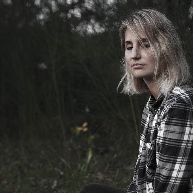 A blonde woman in a flannel shirt gazes downward in a thoughtful pose amidst a forest backdrop.