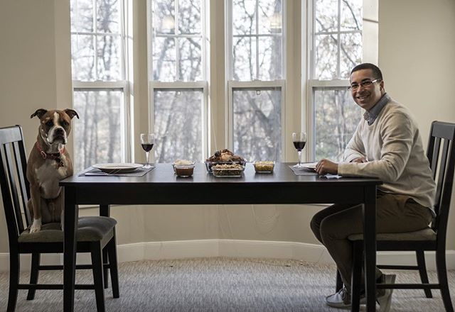 A man and his dog enjoy a dinner together at a table by a bright window.