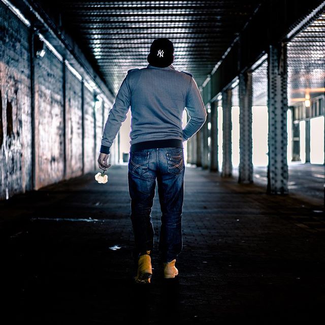 A person in a baseball cap walks through a tunnel holding a flower in a contemplative urban scene.