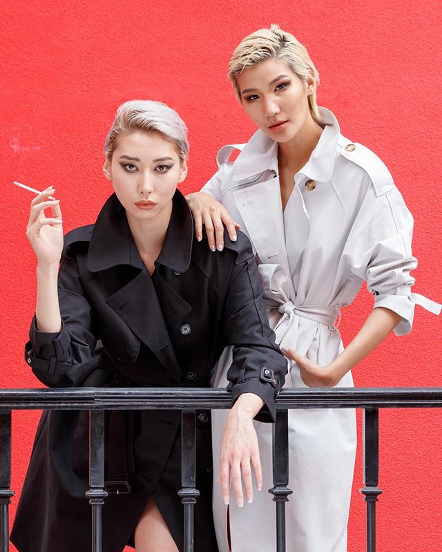 Two fashionable women in trench coats pose against a red wall with serious expressions and stylish aesthetics.