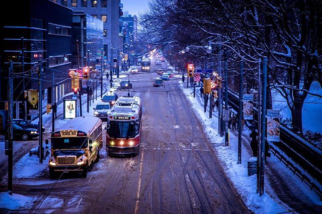 A snow-covered city street with a trolley, school bus, and cars navigating the winter landscape during evening.