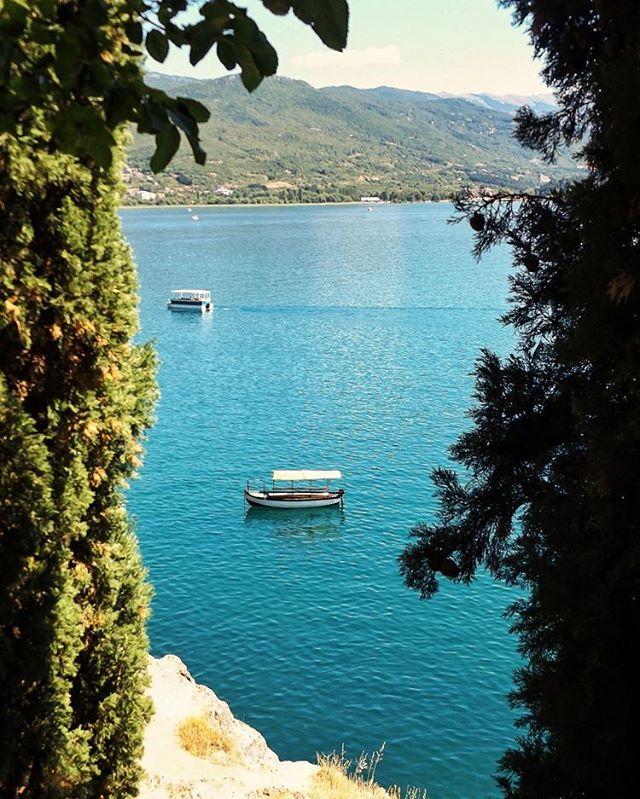 Scenic view of a calm lake with boats, framed by lush trees and majestic mountains in the background. 