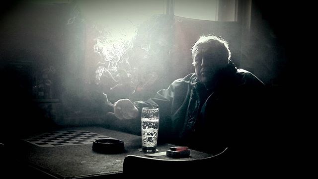 A man smokes a cigarette at a table with a drink in a moody, noir-style scene.