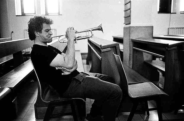 A man sits on a church pew playing a trumpet in a black and white image.