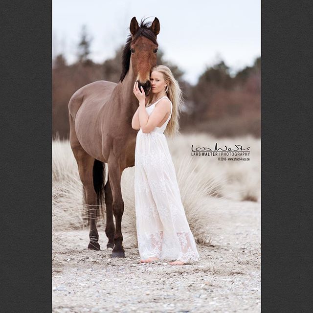 A woman in a white dress stands lovingly with her horse on a beach.