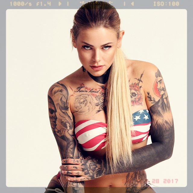 A tattooed woman with blonde hair poses in a studio, showcasing body art and an American flag-themed top.