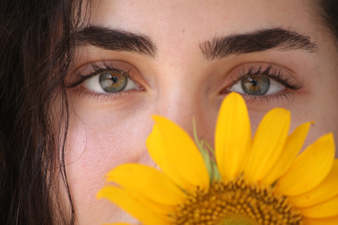 A close up of a woman's face with a sunflower partially covering it, highlighting her eyes.