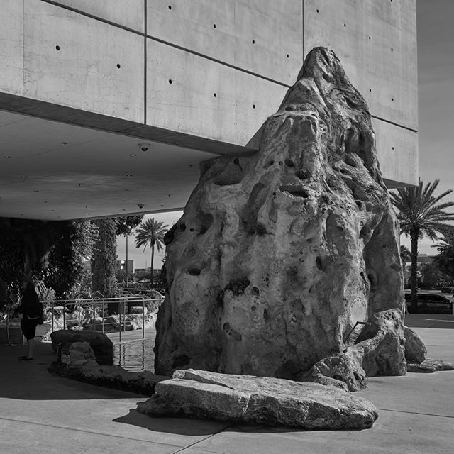 A black and white photo features a rock formation by a modern building with palm trees, creating a unique urban scene.