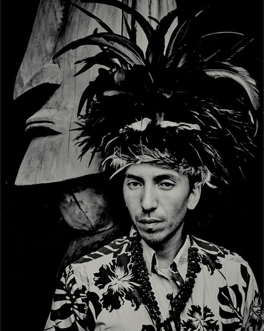A man in a headdress and floral shirt stands in a black and white portrait, showcasing a unique cultural heritage.