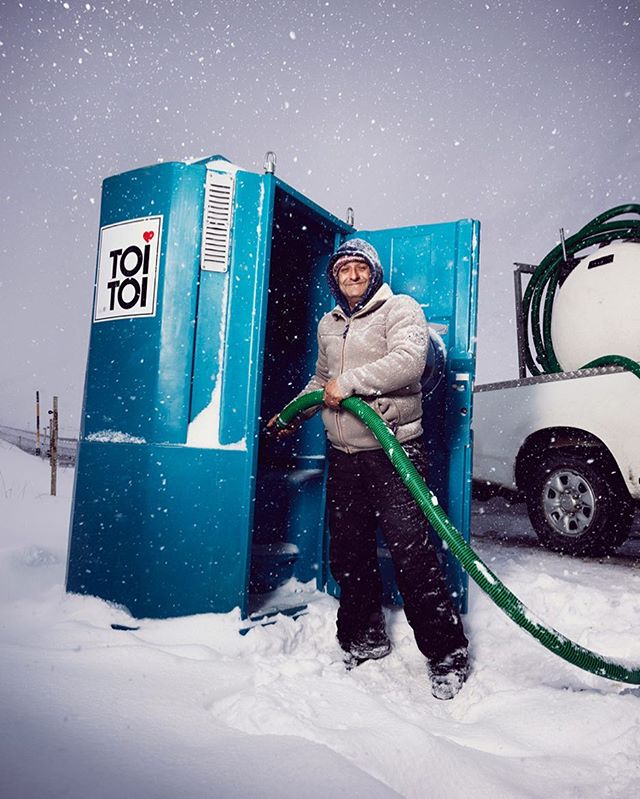 A man stands next to a portable toilet in the snow, holding a green hose, showcasing winter sanitation solutions.