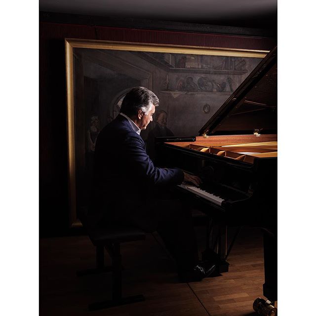 An elegant man plays piano in a warmly lit music room with a classic painting in the background.