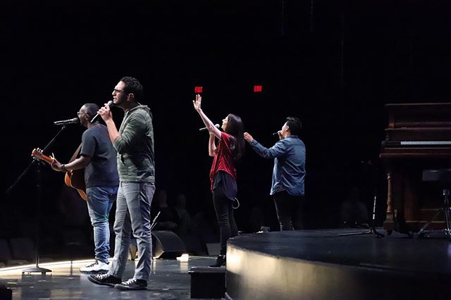 A group of musicians perform on a stage with microphones and instruments in a concert hall setting.