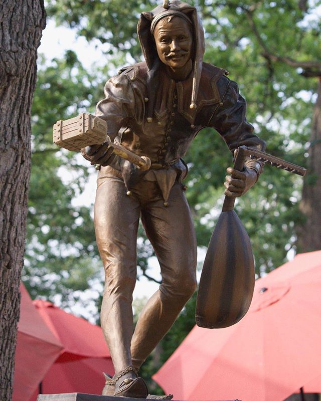 A bronze statue of a jester in full costume, holding props, stands outdoors.