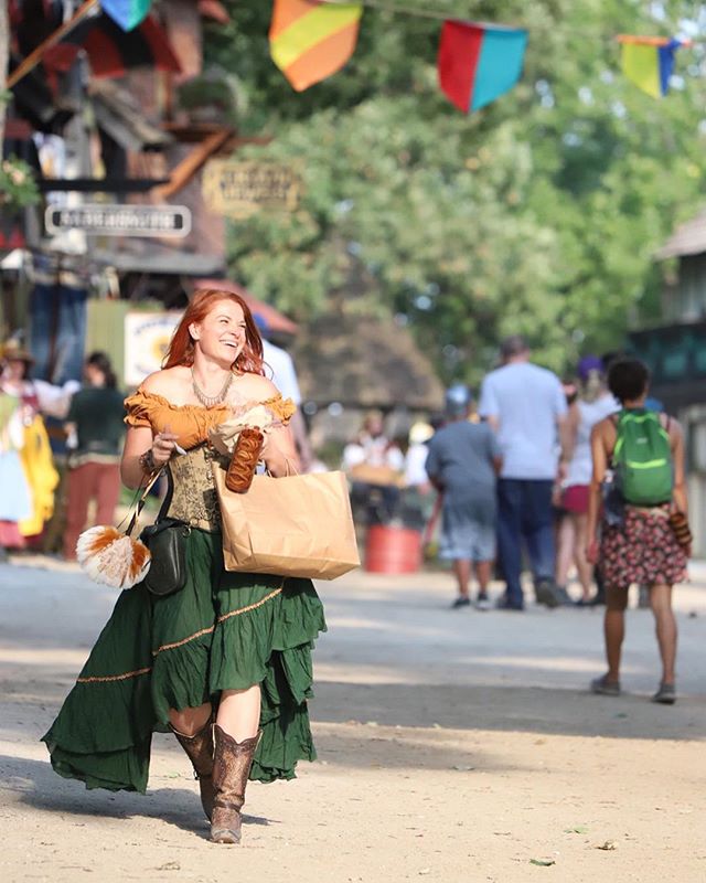 A red-haired woman in renaissance attire walks happily in a fair with a bag and snacks.