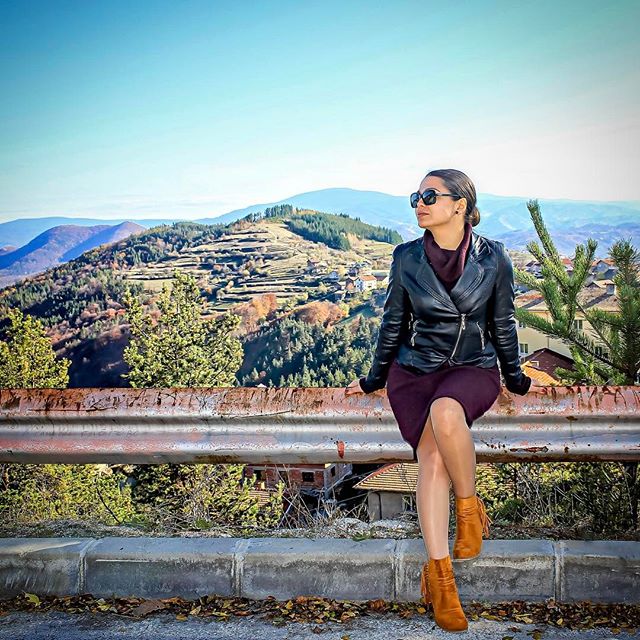 A woman in a leather jacket enjoys the scenic mountain view while sitting on a roadside barrier.