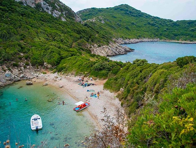 A scenic beach with boats, people swimming, and clear turquoise water surrounded by green hills.
