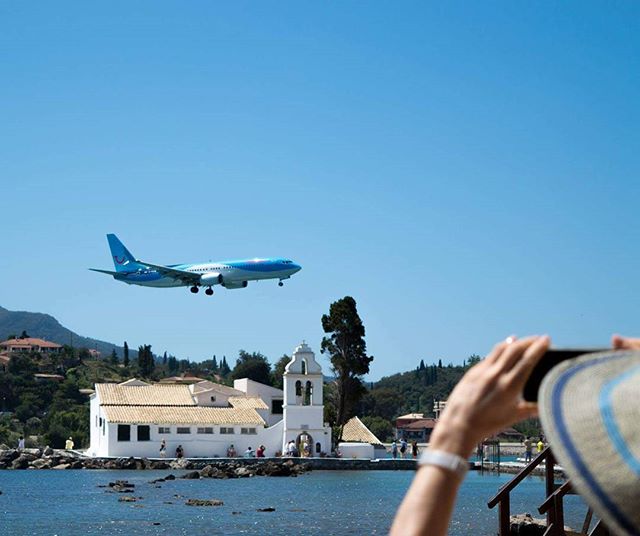 A woman photographs a blue airplane flying over a white church by the sea on a bright day.