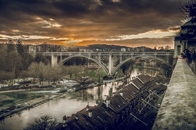 A picturesque cityscape featuring a bridge over a river in Bern, Switzerland, under a moody sky.