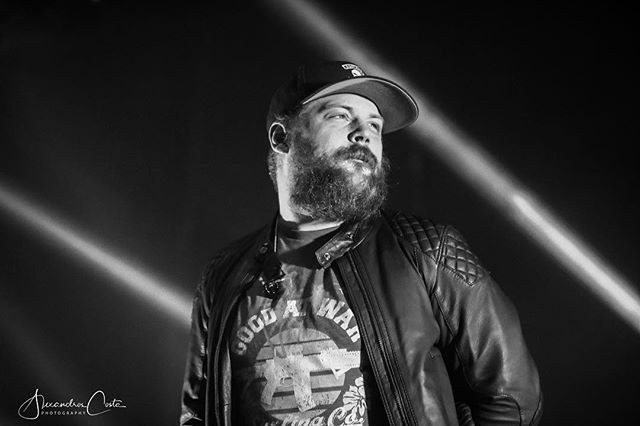 Monochrome shot of a bearded man wearing a cap and leather jacket on stage.