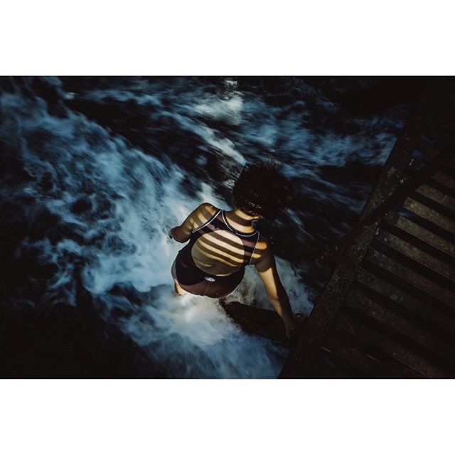 A woman in a swimsuit stands at the edge of a wooden platform next to a flowing waterfall.