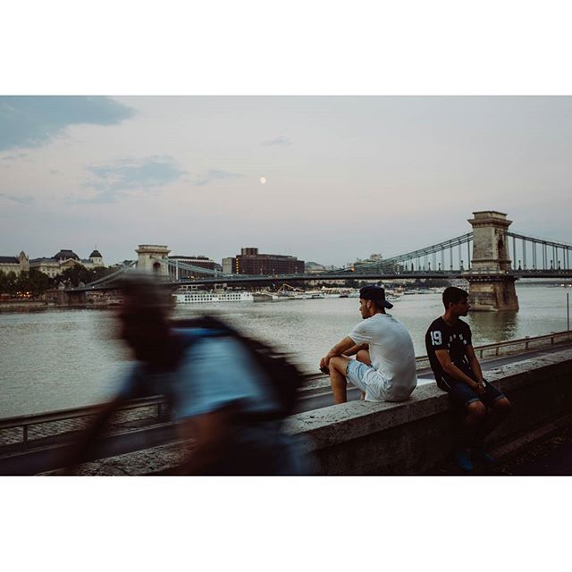 Two young men sit by the river with a view of the Szechenyi Chain Bridge in Budapest at dusk.