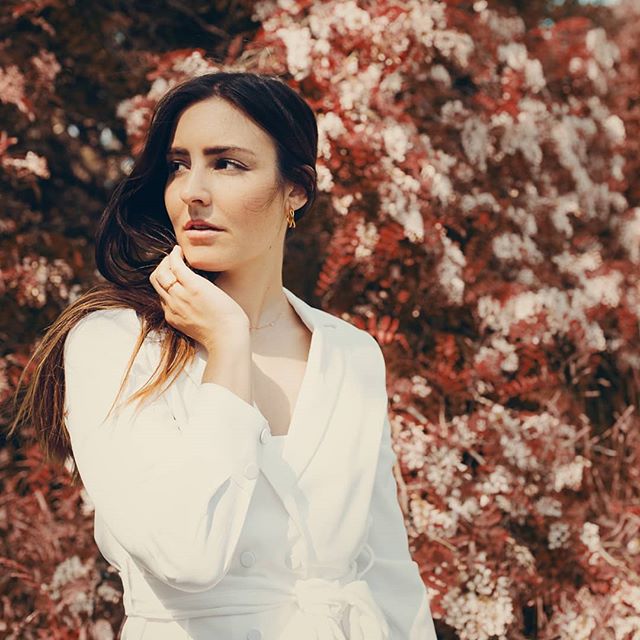 A brunette woman in a white dress poses outdoors with a dreamy expression in a garden setting.