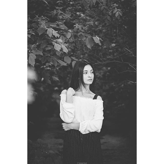 A young woman in a white top stands among foliage in a black and white portrait.