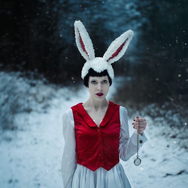 A woman in a white rabbit costume holds a pocket watch in a snowy, dreamy forest setting.