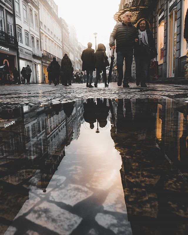 People walk along a wet city street where the buildings reflect in a puddle.
