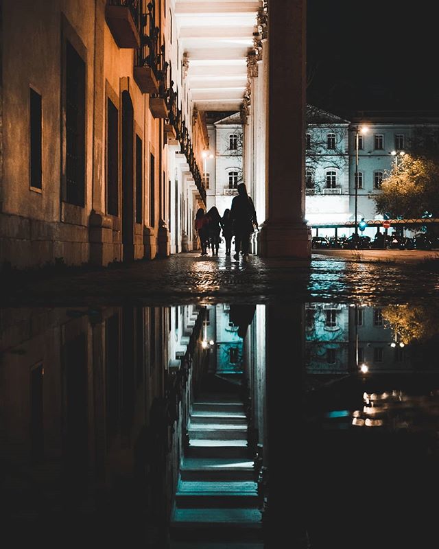 A group of people walks down a wet city street at night, reflected in a puddle.