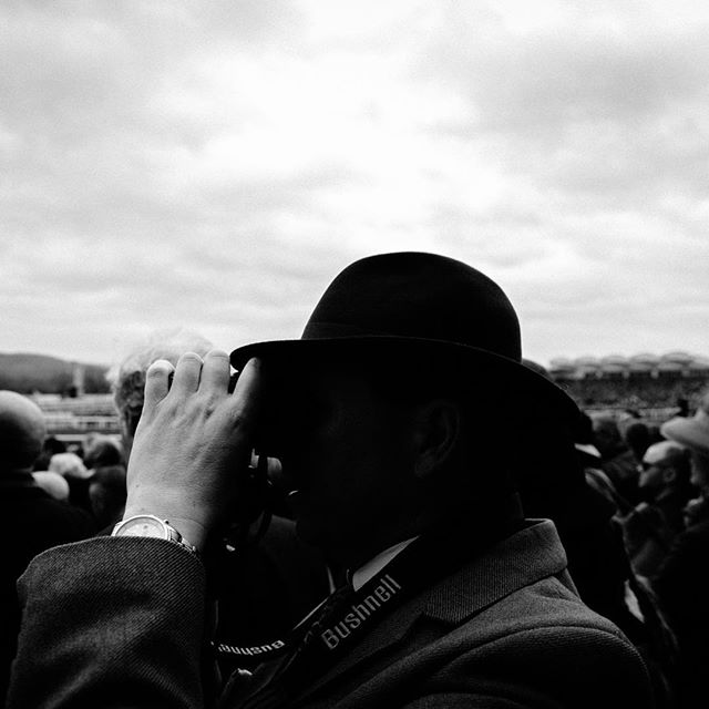 A man in a hat uses binoculars at an event in a black and white photograph.