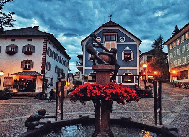 A picturesque town square featuring a fountain and colorful buildings under a cloudy sky.