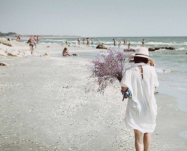 A woman in a white dress walks on a beach, holding a bouquet of purple flowers.