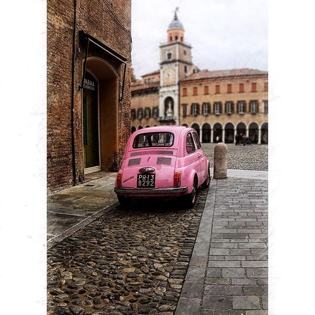 A charming pink vintage car sits on a cobblestone street in an Italian city with historic buildings.