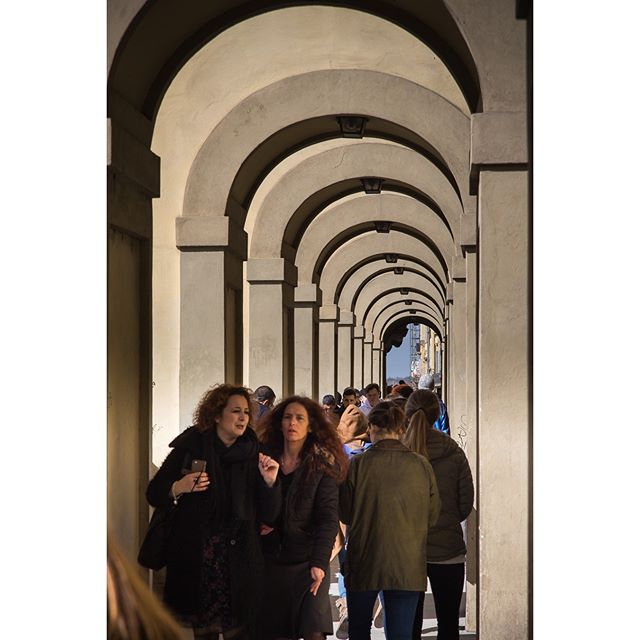 People walk under a colonnade of arches in a European city.