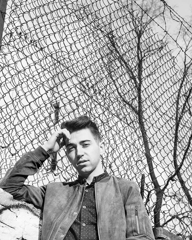 A stylish man poses in black and white against an urban chain-link fence backdrop.