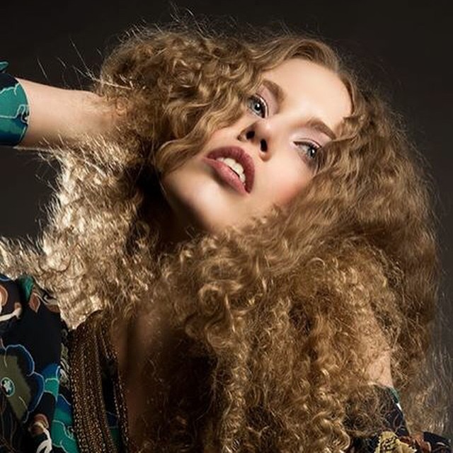 A woman with curly hair poses in a close-up editorial style shot with soft, diffused lighting.