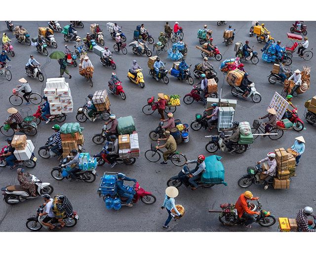 A bustling street scene shows many people transporting goods on motorcycles and bicycles.