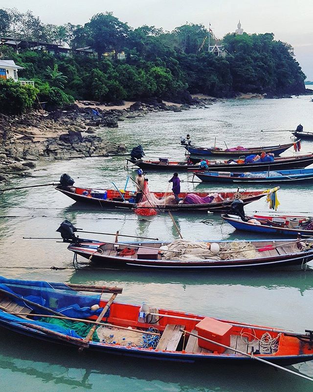 Several fishing boats sit in the tranquil waters of a tropical coastline with a temple visible on the lush, green hillside.