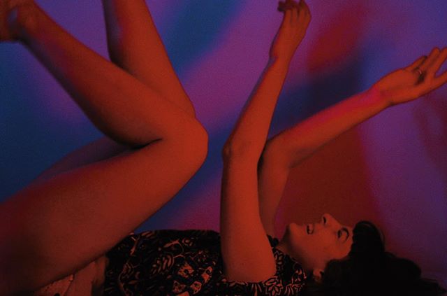 A woman poses with arms raised and legs angled in vibrant red and blue lighting.