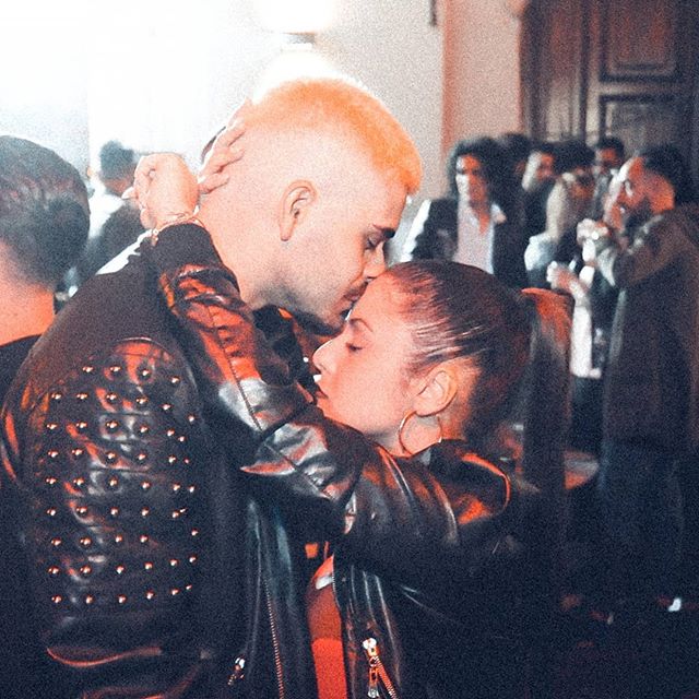 A couple shares an intimate moment at a social gathering, surrounded by other people.