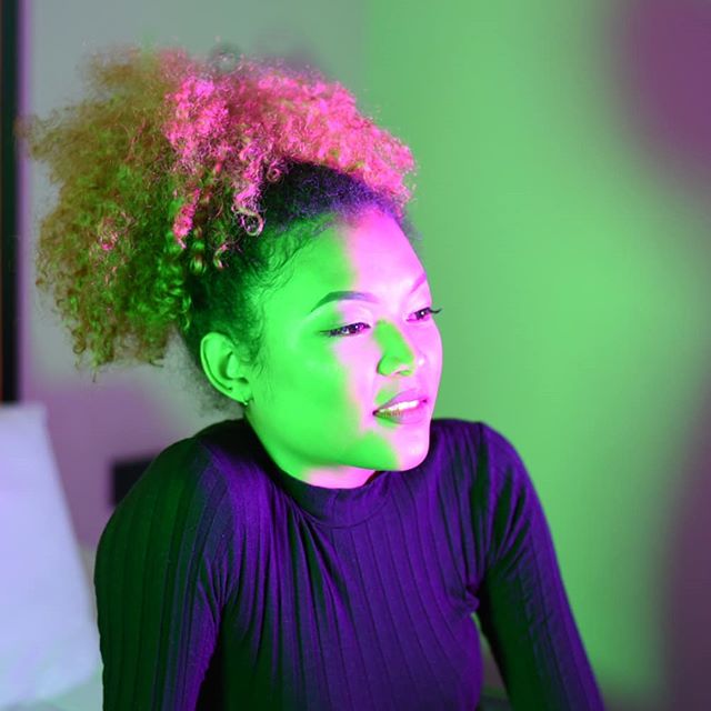A woman with pink curly hair poses in front of a green and purple background.